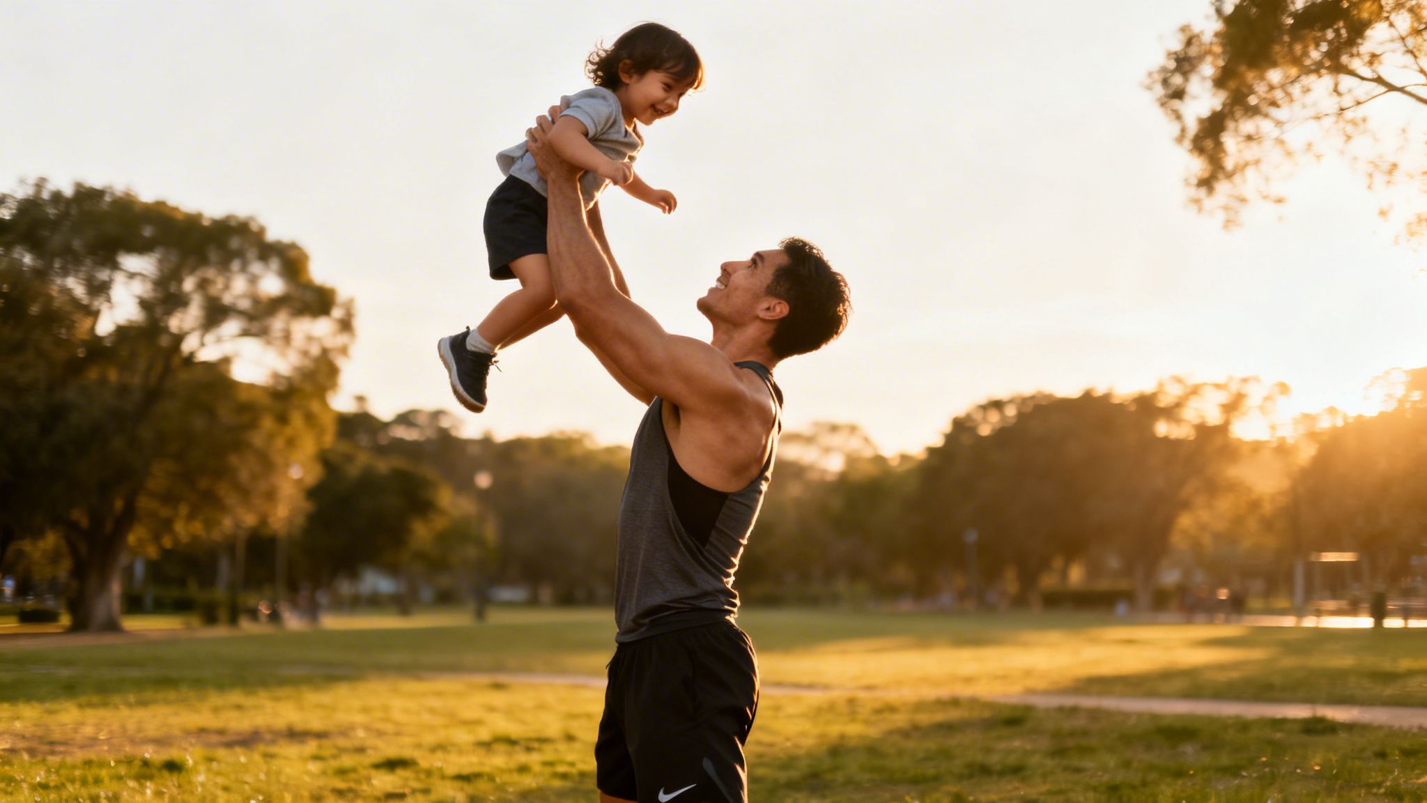 An athletic man holds a smiling child high in the air in a sunlit park.